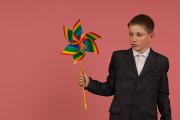 a schoolboy in a black suit with painted nails with a rainbow weather vane in his hand stands on a colored background