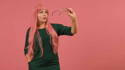young woman with long pink hair in a green dress examines glasses on a pink background