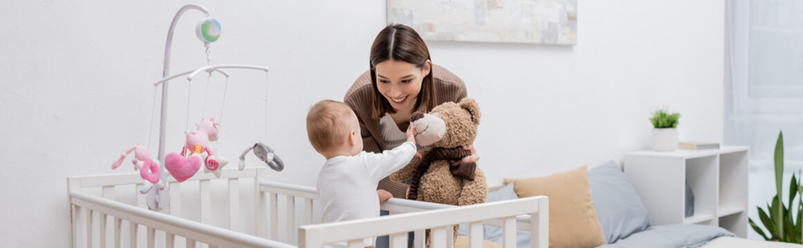 Young mother holding soft toy near baby boy in crib, banner.