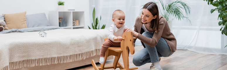 Smiling baby boy sitting on rocking horse near parent in bedroom, banner.
