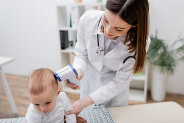 Young family doctor holding pyrometer near head of baby boy in clinic.