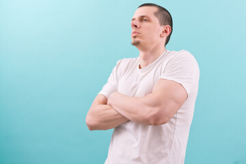 A caucasian strong man in a white t-shirt is standing confidently with crossed hands on a blue background. Person. Male. Man. Guy. Model. Studio. Young. Standing. Portrait. Attractive. Casual. Hand