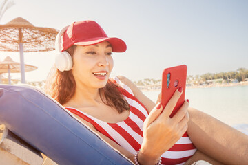 Happy girl is lying on a sunbed, sunbathing and listening to music or a podcast using headphones...