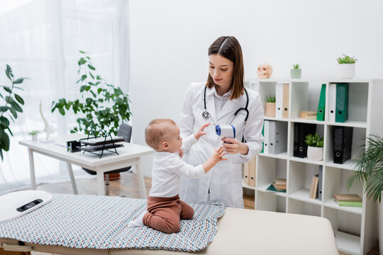Young family doctor holding non contact pyrometer near baby on medical couch in clinic.