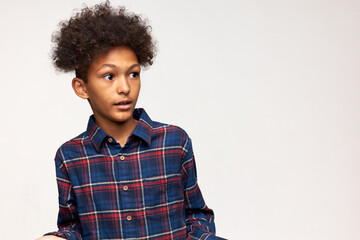 Horizontal portrait of teen African boy in flannel shirt with afro haircut standing against white background looking aside with puzzled and astonished face expression with fear in his eyes