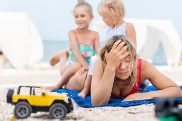 Fototapeta na wymiar Two cute little sibling girls enjoy having fun playing sitting on tired exhausted mother's back at sea ocean beach. Frustrated mom make face palm gesture. Vacation family small kids trouble concept
