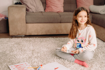 A little beautiful girl in casual clothes sitting on the carpet at home holding a book in her hands and looking aside with smile.