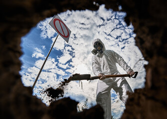 View inside pit of scientist in white suit and gas mask standing near prohibition sign and digging...