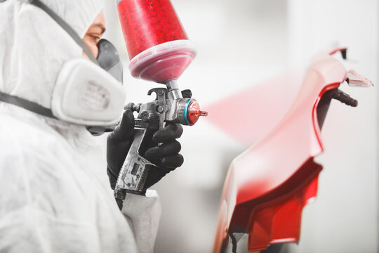 Auto mechanic worker painting car fender with spray gun in a paint chamber during repair work.