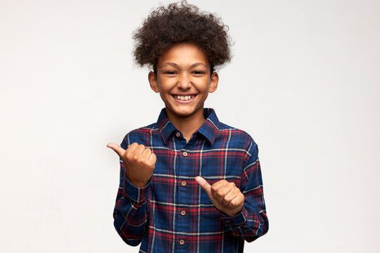 Cheerful laughing charismatic dark-skinned boy standing against white studio wall with blank copy space for your advertising content keeping thumbs up pointing them to the right, dressed in shirt