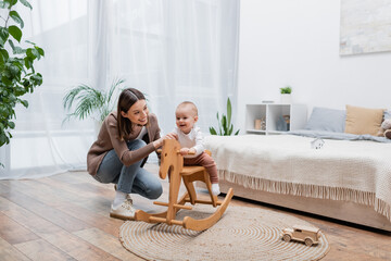 Positive baby boy sitting on rocking horse near mom in bedroom.