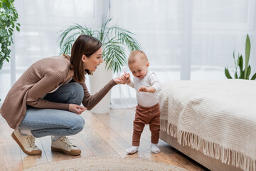 Obraz premium Happy mother holding hand of smiling baby near bed at home.