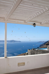 A whitewashed terrace overlooking a crowd of seagulls flying around a swimming pool and the aegean sea in the background in Santorini