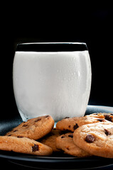 A glass of cold milk and chocolate chip cookies on a dark plate band a black background, vertical macro photography