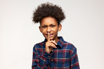 Fototapeta na wymiar Be quiet. Handsome beautiful African boy with afro hair dressed in plaid shirt looking at camera with frustrated face expression, putting index finger next to his mouth, asking for silence