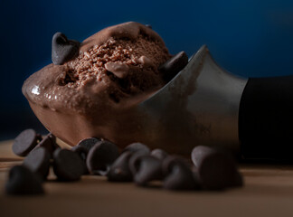 An ice cream spoon with melted chocolate ice cream, and chocolate chips on the side, and a glass with ice cream in the background with dark blue background.