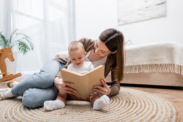Young mom reading book with baby son on floor in bedroom.