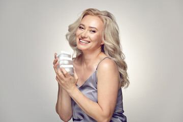 Senior blonde woman holding multiple jars of cream for face moisturising on grey isolated.