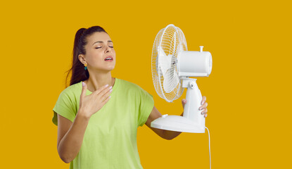 Young woman with a fan in her hand is exhausted from the heat, trying to cool off by the fan, oppressive heat, too hot weather and sultriness in summer, flat without air-conditioner concept. - obrazy, fototapety, plakaty