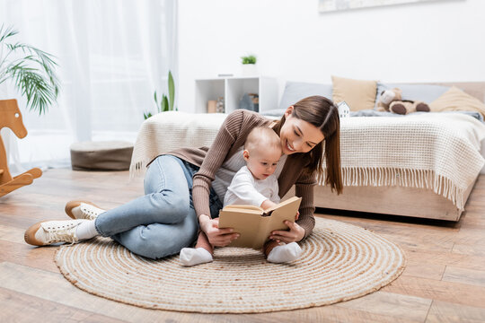Smiling parent reading book near baby boy on rug at home.