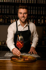 smiling male bartender holds glass with signature cocktail in his hand and looks at camera
