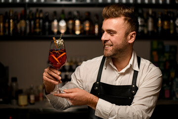 smiling male bartender holds glass with signature cocktail in his hands and looks at it