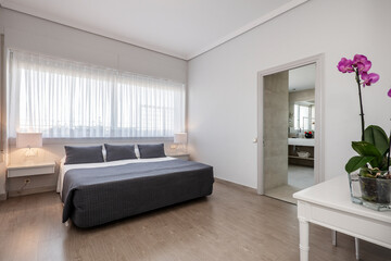 Bedroom with a large double bed with gray cushions to match the bedspread, a white wooden desk, twin lamps on the bedside tables and a large window above the bed