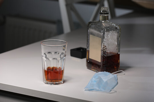 Glass With Alcohol, Bottle Of Whiskey And Medical Mask On The Table. Concept Of Cancelled Social Events During Pandemic And Corona Virus Quarantine