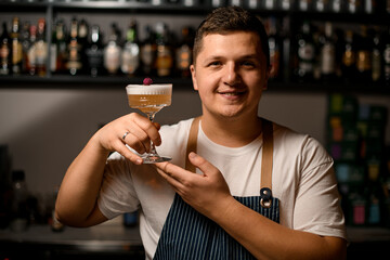 smiling bartender holds crystal wine glass with tasty foamy cocktail in his hands.