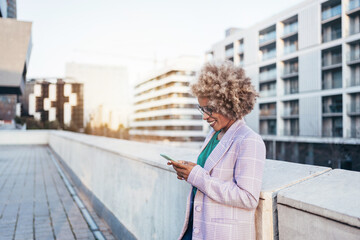 Smiling African American business woman with afro hair using smartphone standing on rooftop at sunset 