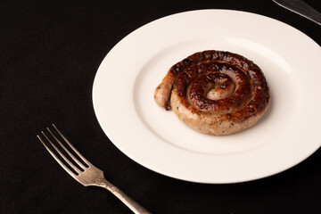 Fried spiral sausage on a white plate with a fork and knife on a black tablecloth
