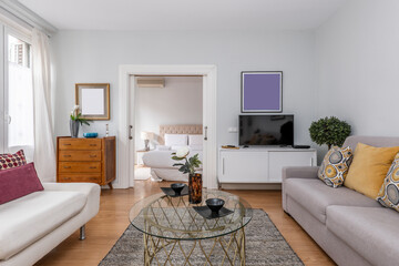 living room with sofas upholstered in fabric, wooden sideboard with flat screen tv, round glass coffee table and entrance to a bedroom in the background