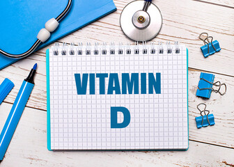 On a light wooden background, a stethoscope, a blue notepad, blue paper clips, a blue marker and a sheet of paper with the text VITAMIN D. Medical concept