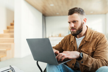 Serious Freelancer Man Using Laptop Computer Wearing Earbuds At Home