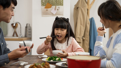 happy Asian family of three eating together in the dining room at home. the girl drops her food as she picks up vegetable to eat