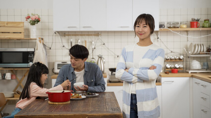portrait of pretty smiling taiwanese mother looking at camera with folded arm as the father and schoolgirl daughter are enjoying meal in the dining room at background