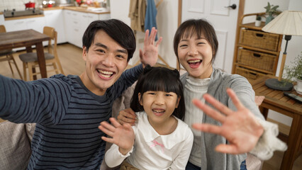 mobile cam view cheerful asian family of three waving hands to camera and talking with smile. they cuddle with each other on the couch in the living room at home