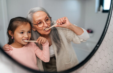 Senior grandmother and granddaughter standing indoors in bathroom, brushing teeth in morning. - Powered by Adobe