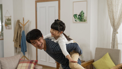 Happy Asian daughter sitting playing on her father’s back at home. she opens arms and acts like...