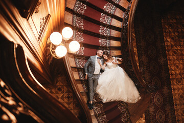 wedding day. beautiful bride in white long dress and young groom wearing in black suit are hugging and lying on the stairs inside big historic europe building
