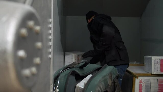 A bank robber clearing out a safe. The burglar is loading bundles of cash into a back inside a bank vault.