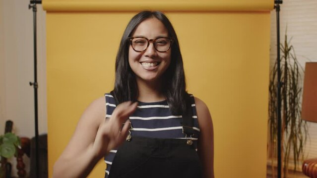 Happy young Asian woman holds peace sign up to the camera in a yellow studio
