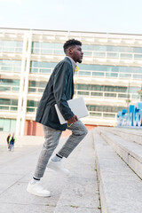 businessman walking up the stairs with a laptop in his hand and an office building behind him.