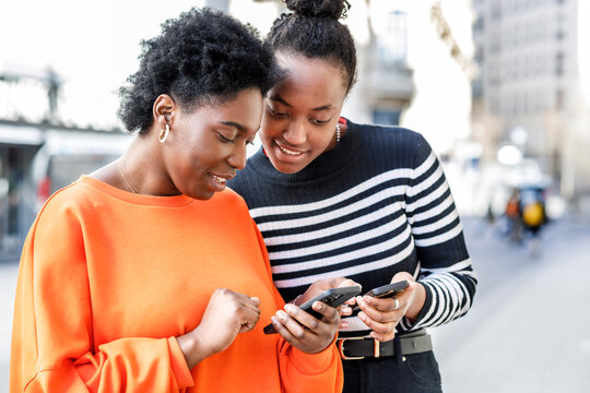 Cheerful black women browsing smartphones on street