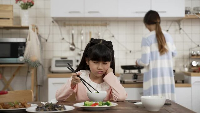 selective focus of unhappy picky Asian schoolgirl daughter holding face with disgust look on face while playing with food in bowl at dining table. her mother is cooking in kitchen at background