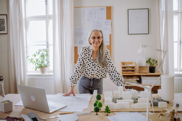 Mature woman architect with model of houses standing in office and looking at camera.