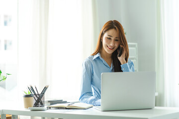 Happy Asian businesswoman sitting at desk behind her laptop and talking with somebody on her mobile phone.