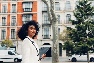 Happy businesswoman walking around city as she carries folder in her hands and looks away