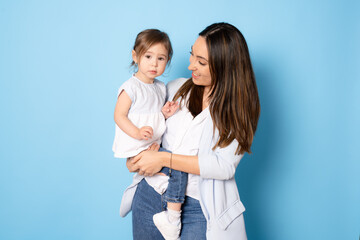 Photo of pretty young mommy hugging little daughter standing isolated over blue background. Motherhood concept.