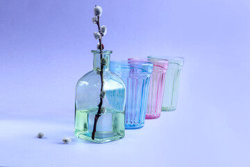 Spring mood: flowering willow branch in a green bottle against the background of multi-colored glasses, blue background, space for text